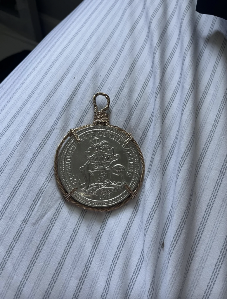 A silver coin encased in an intricate wire wrap lies on a white and gray striped fabric. The coin features detailed engravings and a coat of arms with the phrase "FORTITUDO ET FIDELITAS.