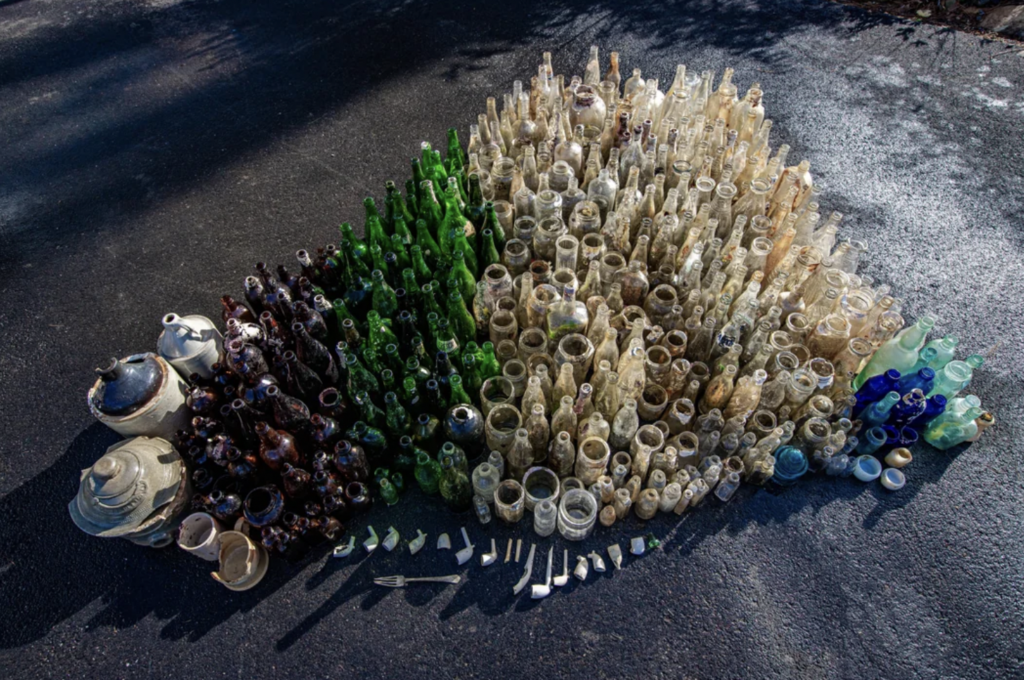 A heart-shaped arrangement of various old bottles and ceramic pieces on a dark, paved surface. The bottles vary in color, including clear, green, and brown hues, with some showing signs of dirt or erosion, suggesting they have been recently unearthed or collected.