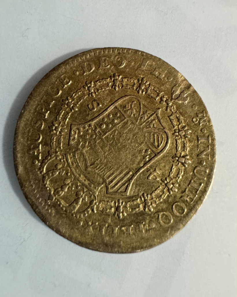 A worn, round coin with a detailed design featuring a shield-like coat of arms in the center surrounded by text and decorative elements around the edges. The coin appears to have historical and possibly antique significance due to its intricate design and patina.