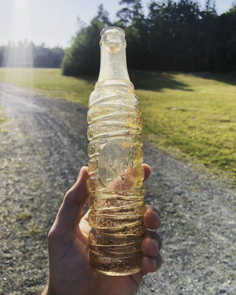 A hand holds an old, clear glass soda bottle with spiral ridges against a backdrop of a gravel path, sunlit grassy area, and trees in the distance. The bottle has some dirt and debris on it, indicating it was found outdoors.