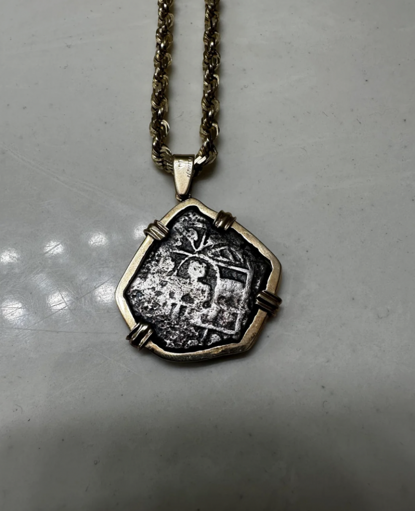 A weathered, silver-colored pendant with an intricate, ancient design hangs from a textured, gold-colored chain. The pendant has a rough, irregular shape with metal prongs securing it. The background is a smooth, light gray surface.