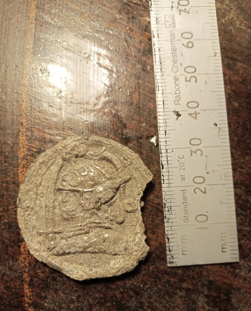 A coin or medallion with a worn depiction of a helmeted figure is placed next to a ruler showing metric measurements. The coin has an irregular, rugged edge. The surface it rests on appears to be wooden and worn.