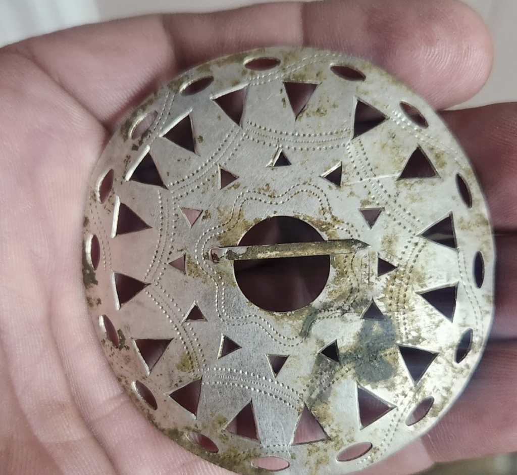 A hand holding a circular metallic object with intricate geometric and floral cut-out designs. The metal appears slightly tarnished with a pin mechanism in the center, indicating it may be a brooch or similar decorative item.