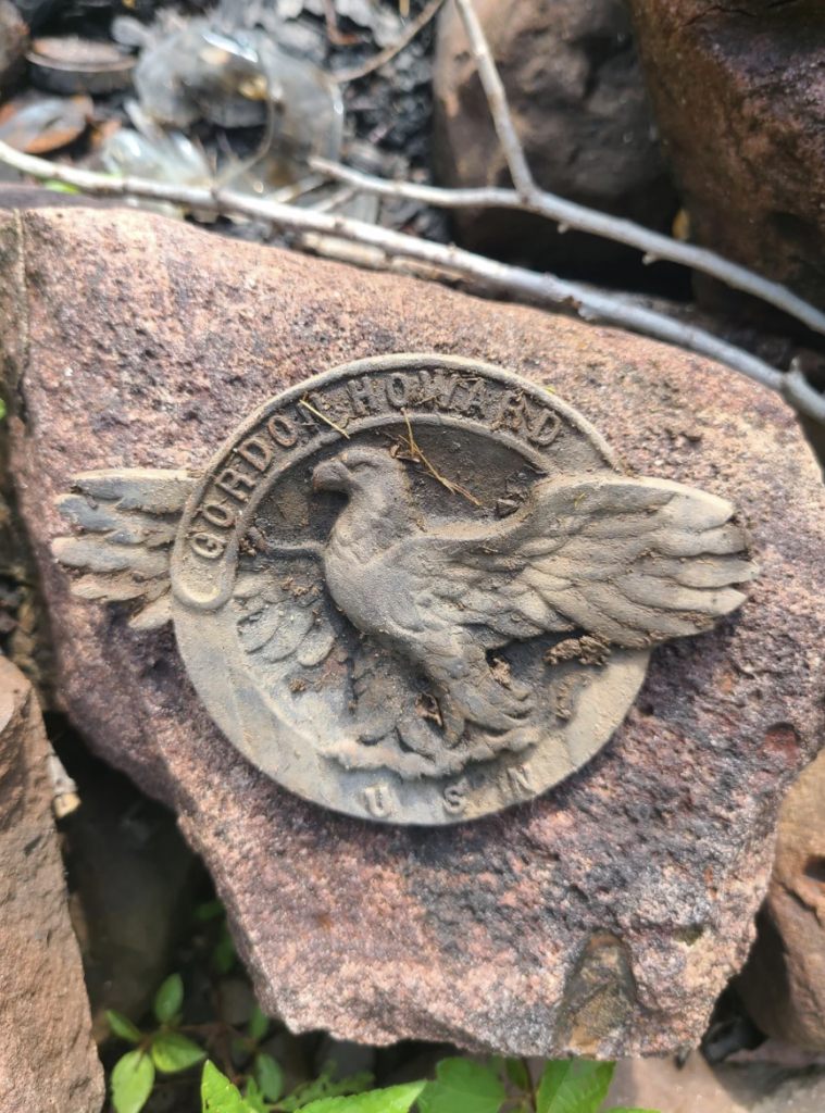 A weathered metal medallion featuring a detailed bird with outstretched wings and the inscription "GORDON HOWARD U.S.W" is partially buried in a rock surface. Surrounding stones and some foliage are visible in the background.