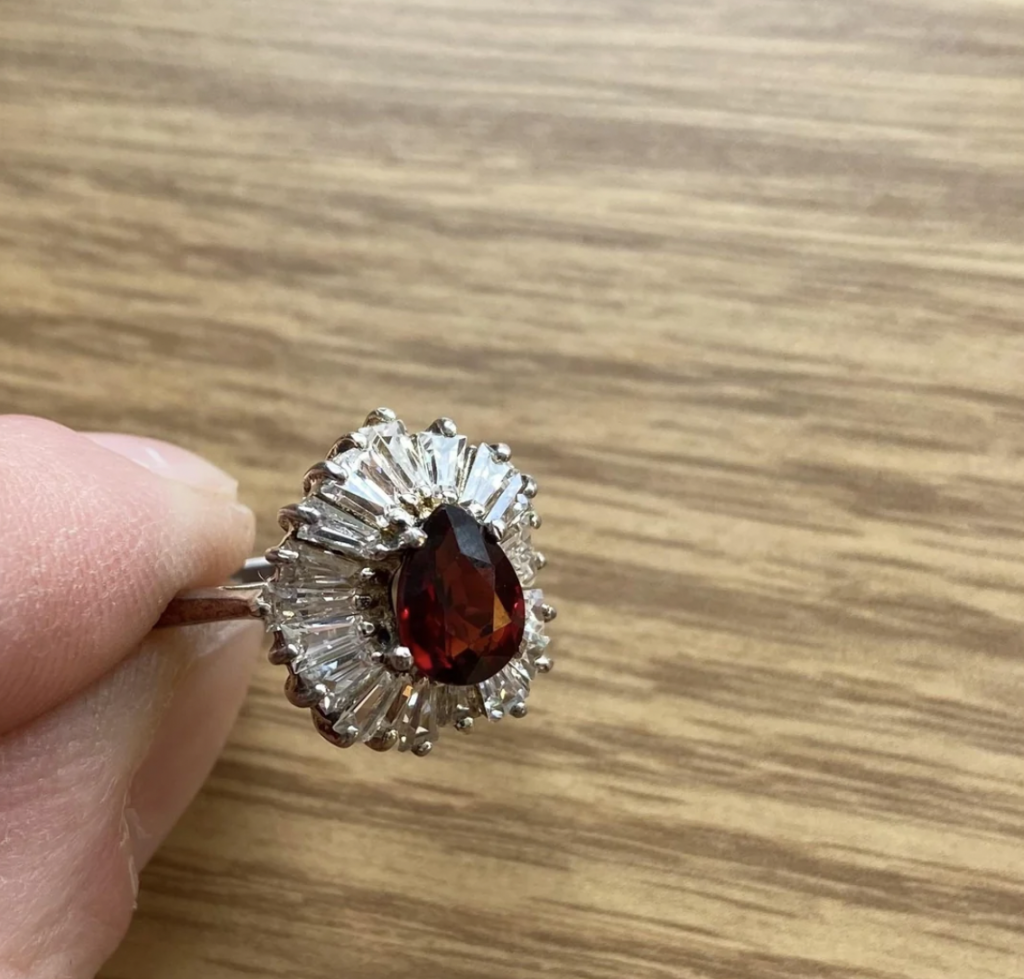 A hand holding a silver ring with a tear-shaped, deep red gemstone in the center. The gemstone is surrounded by a halo of small, clear, baguette-cut stones. The background is a wooden surface.