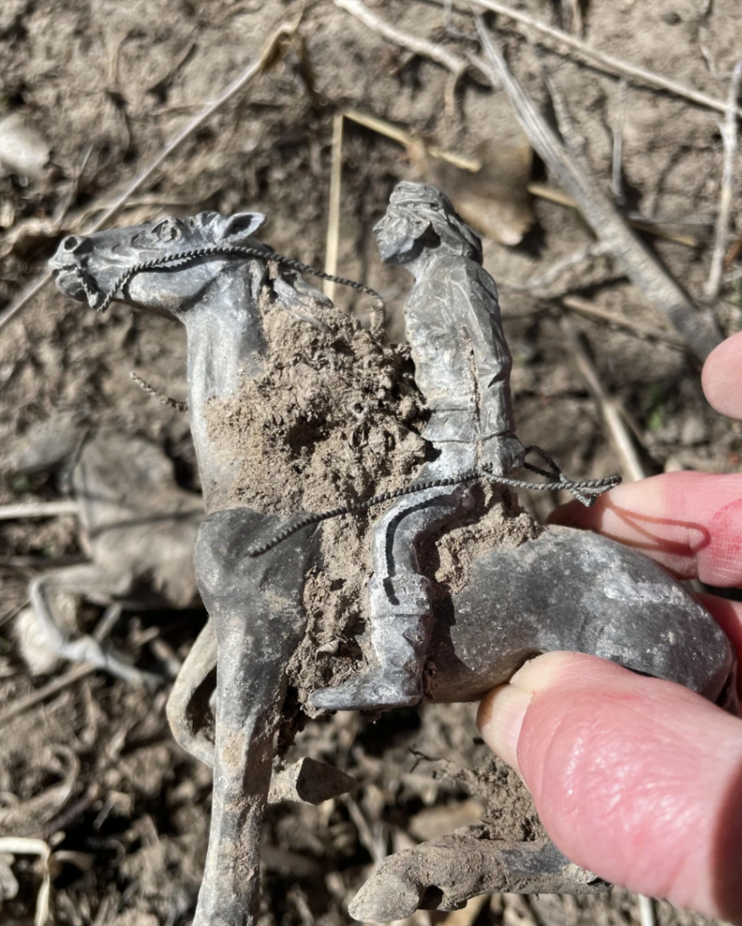 A hand holding a small, dirt-covered toy figure of a person riding a horse. The toy appears to be metallic and is partially buried in the dirt. Dry grass and twigs can be seen in the background.