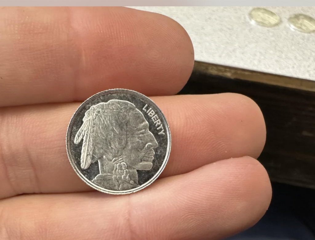 Close-up of a hand holding a coin featuring a Native American profile on one side with the word "LIBERTY" inscribed. Other coins are slightly visible in the blurred background to the right.