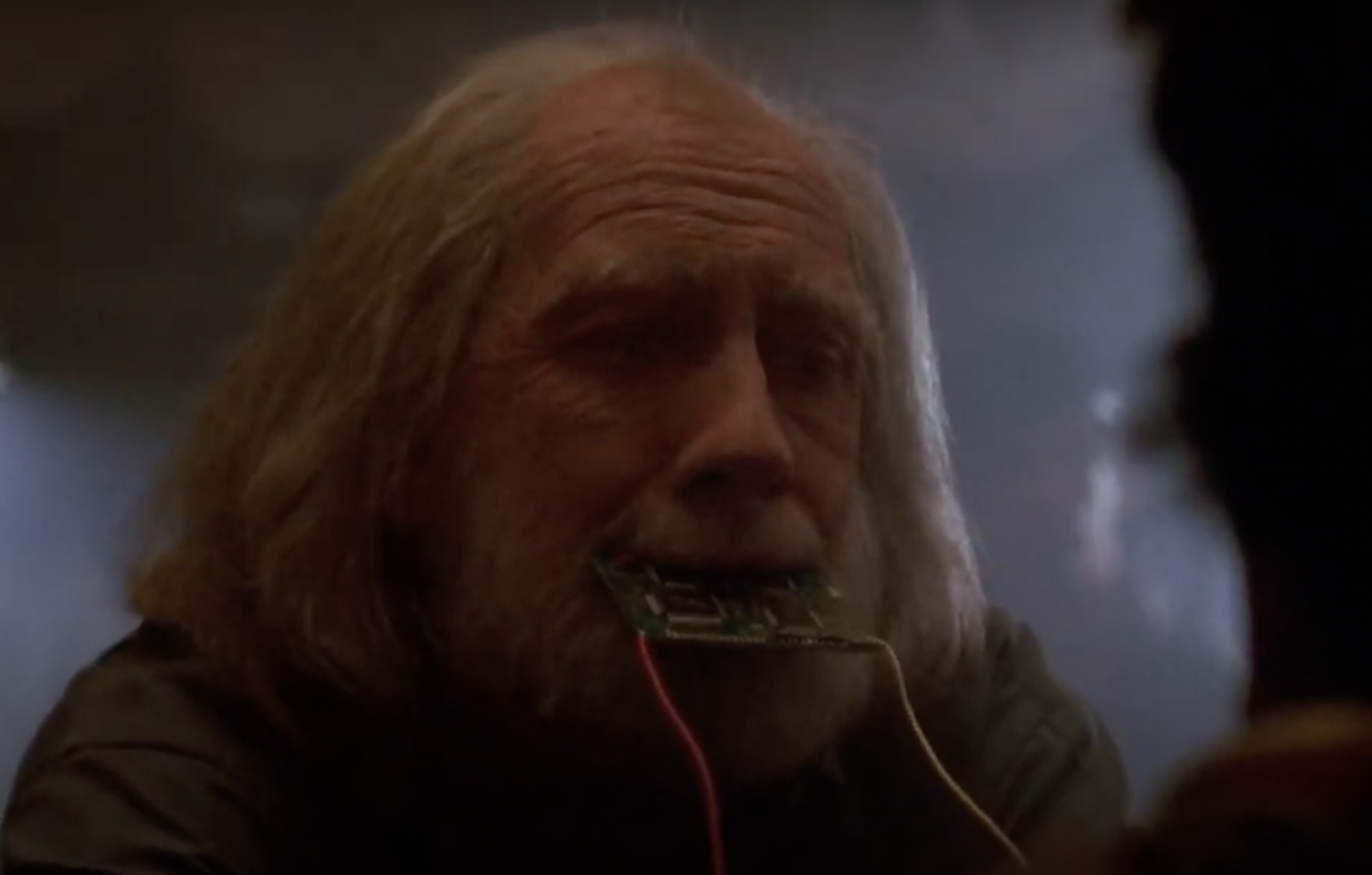 A man with long white hair and a beard has a circuit board with attached wires protruding from his mouth. The background is dimly lit, creating a mysterious and intense atmosphere. The man looks distressed or in pain.