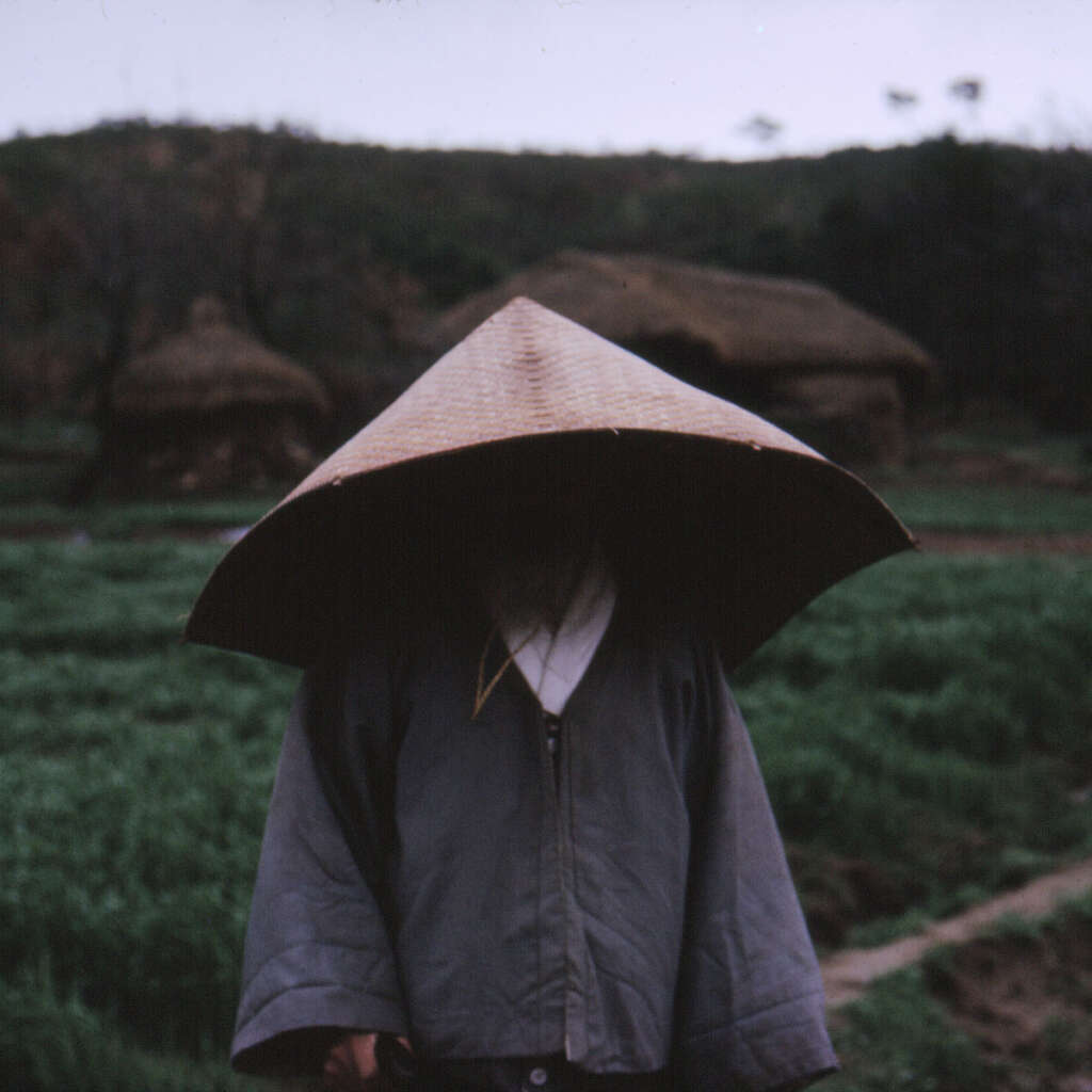 A person stands facing the camera wearing a large conical woven hat that obscures their face, a dark robe, and a light-colored undergarment. The background is a rural landscape with blurred, thatched-roof structures and greenery visible.