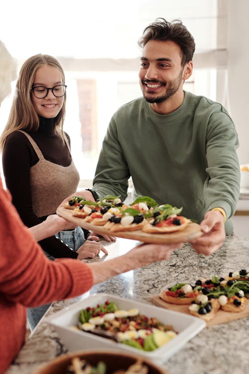 A man in a green sweatshirt and a woman in a brown sweater with glasses smile as they pass dishes of vegetable-topped flatbreads across a kitchen island. Fresh salads and more flatbreads are visible on the countertop.