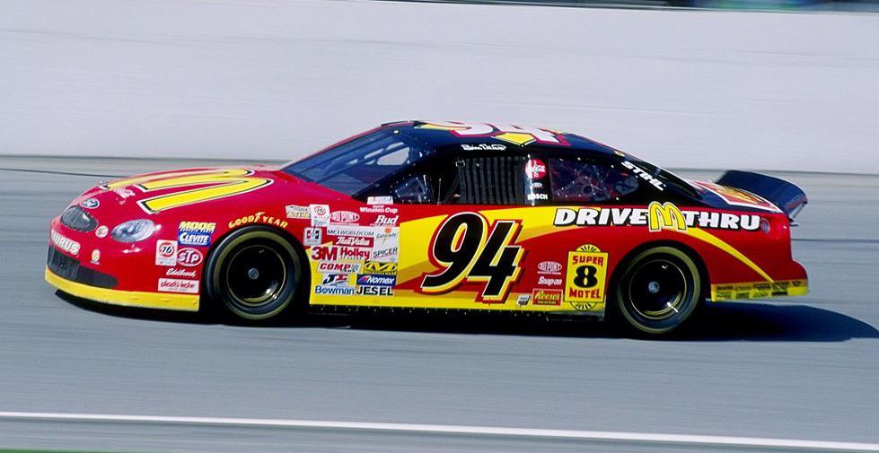 A red and yellow stock car numbered 94 speeds along a racetrack. The car features various sponsor logos, including a prominent fast food chain logo on the hood and the side. The driver is visible through the window, wearing a helmet and racing gear.