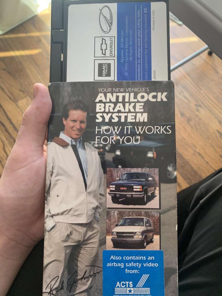 A person is holding a manual labeled "Your New Vehicle's Antilock Brake System: How It Works For You" featuring a man in a white jacket, and images of a black vehicle. An airbag safety video tape is partially inserted in a device at the top.