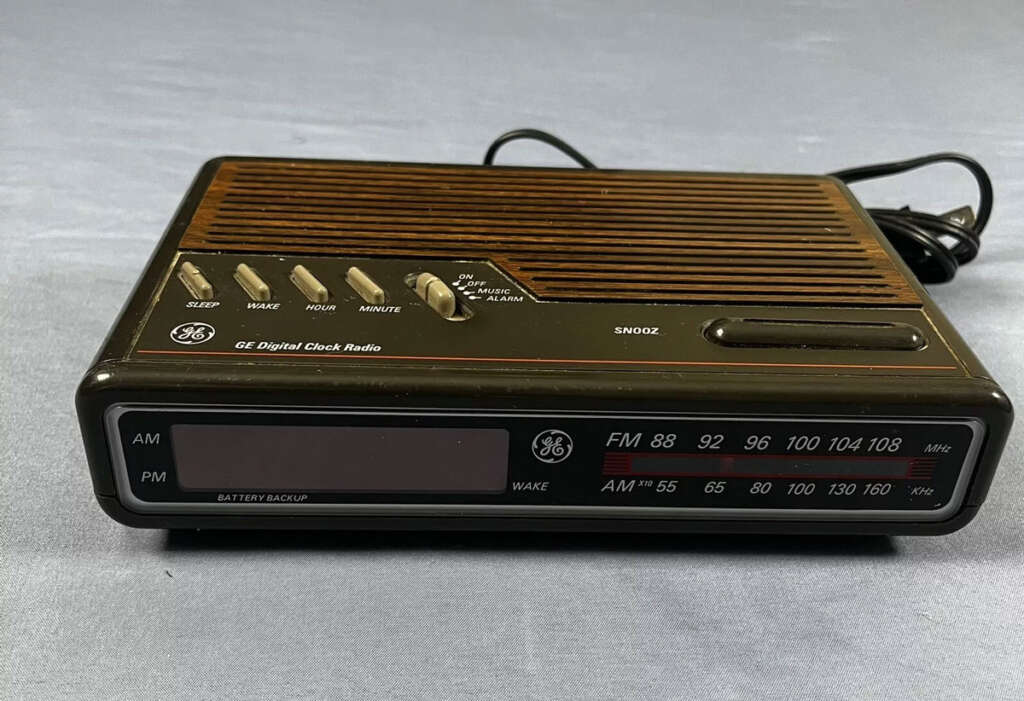 A vintage GE digital clock radio with a wood-grain finish and black casing. It features a digital display for AM and FM radio frequencies, buttons for tuning, an alarm, and sleep functions, and a battery backup indicator. The device has an attached power cord.