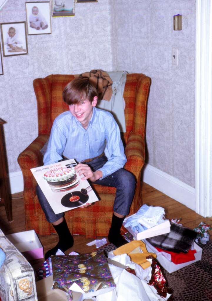 A young boy sits in a plaid armchair, smiling while holding a vinyl record album. Wrapping paper and opened presents, including clothes, surround him on the floor. Framed pictures decorate the wall behind him, and a coat hangs from the back of the chair.