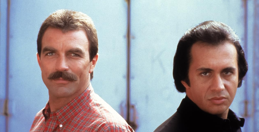 Two men stand back-to-back against a light blue background. The man on the left has a thick mustache and is wearing a red plaid shirt. The man on the right has dark, slicked-back hair and is dressed in a black jacket. Both are looking directly at the camera.