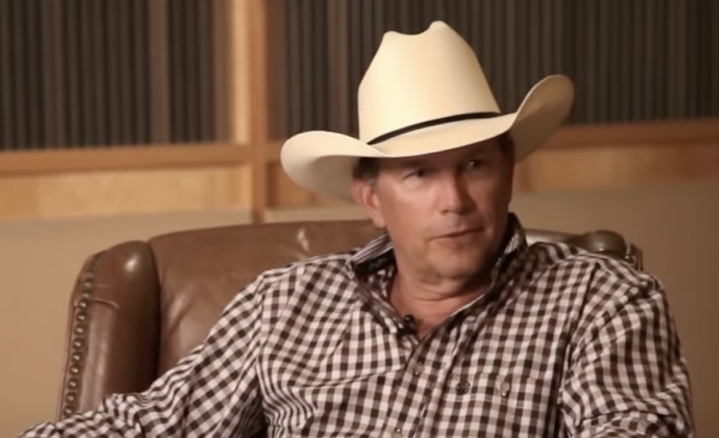 A man wearing a white cowboy hat and a black and white checkered shirt is sitting on a leather chair. He is looking slightly to his right. The background features wooden paneling and a gray wall.