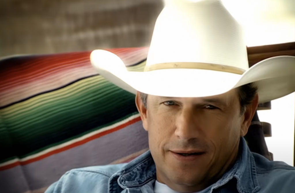 A man wearing a white cowboy hat and blue denim shirt is sitting in front of a colorful striped blanket. He is looking directly at the camera and appears to be outdoors, with a blurred background. He has a slight smile on his face.