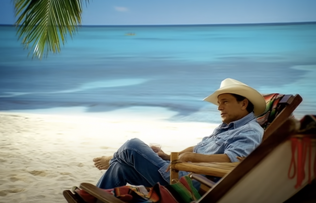 A person wearing a cowboy hat and jeans sits in a colorful beach chair on a sandy beach. They are looking out over the calm, turquoise ocean, with a palm frond partially visible in the upper left corner of the image. The sky is clear and blue.