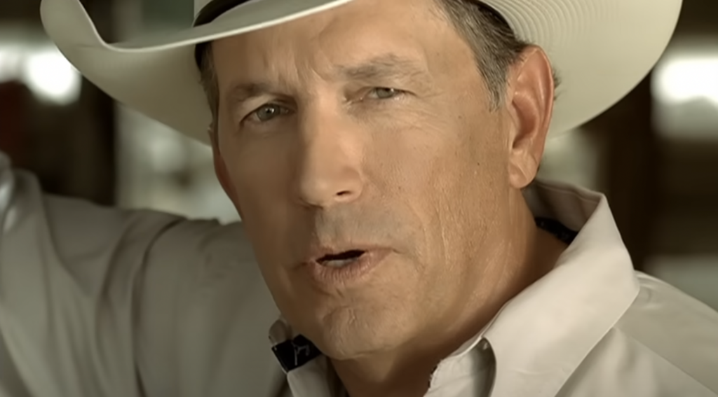 A middle-aged man wearing a white cowboy hat and a light-colored button-up shirt is looking into the camera with a serious expression. The background is blurred, focusing attention on the man's face.