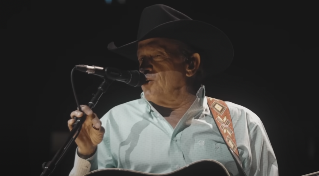 A man in a black cowboy hat and light blue shirt sings into a microphone while playing an acoustic guitar. He is illuminated by stage lights against a dark background.