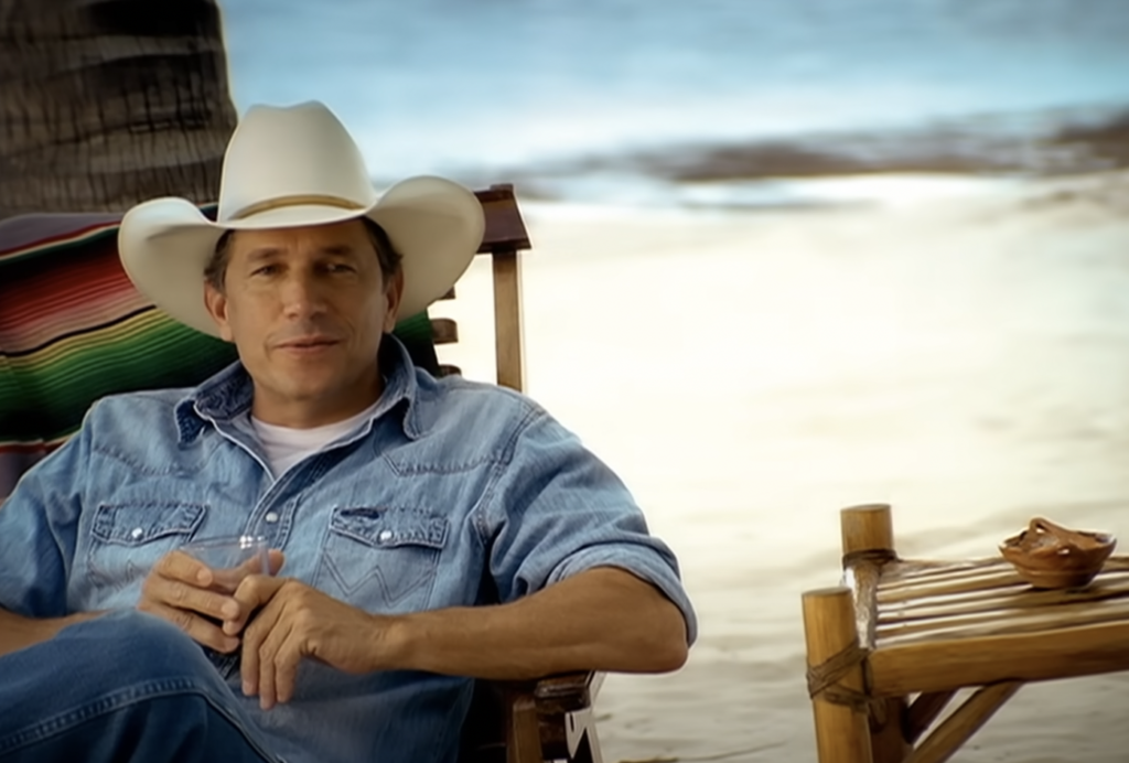 A man wearing a white cowboy hat and denim shirt sits in a wooden chair on a sandy beach. A colorful blanket is draped over the chair. A small table with a ceramic dish is beside him, and the ocean is blurred in the background.