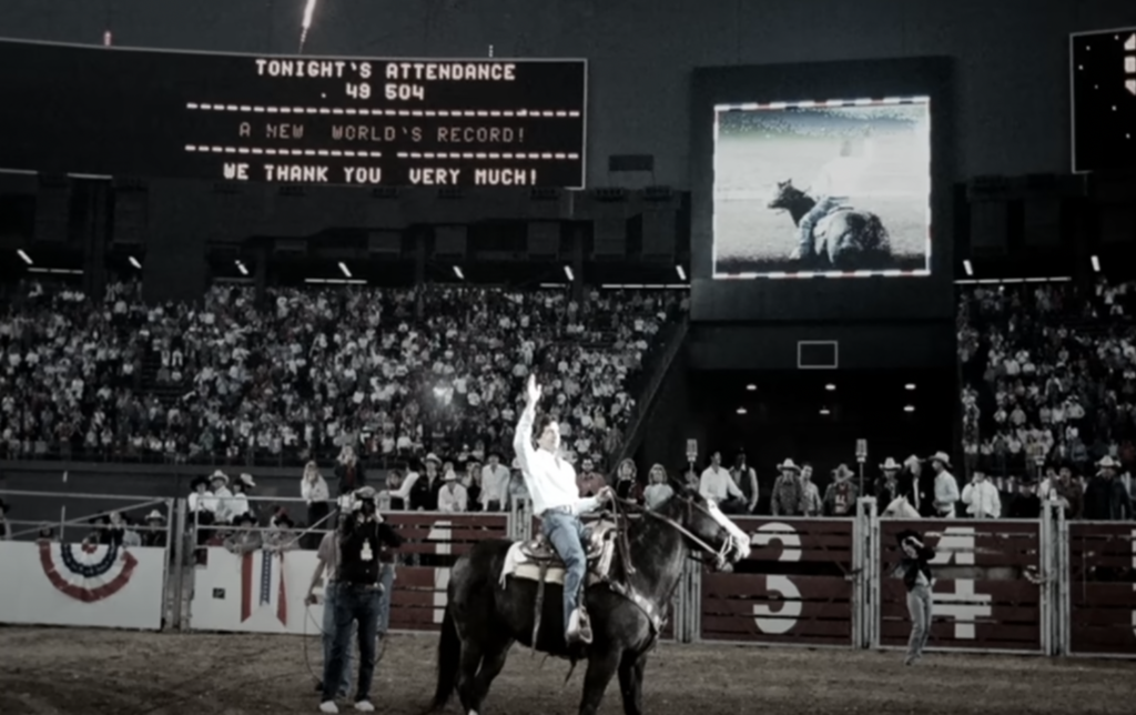 A rodeo rider on a horse raises their arm in celebration in front of a large, applauding stadium crowd. A scoreboard behind displays "Tonight's Attendance 48,504" and "A New World's Record!" Additionally, a giant screen shows a bull rider in action.