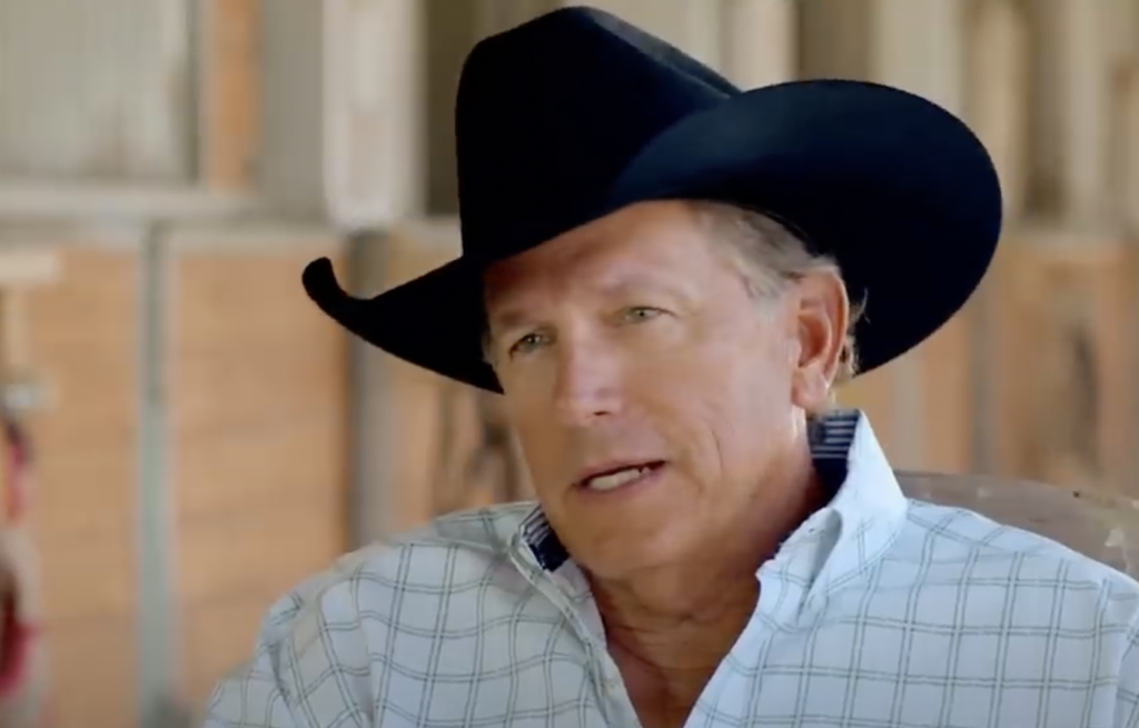 A middle-aged man wearing a black cowboy hat and plaid shirt is talking, likely in a rural or Western setting. He has light-colored hair and appears to be speaking with a relaxed expression. The background is slightly blurred, suggesting an indoor or barn setting.