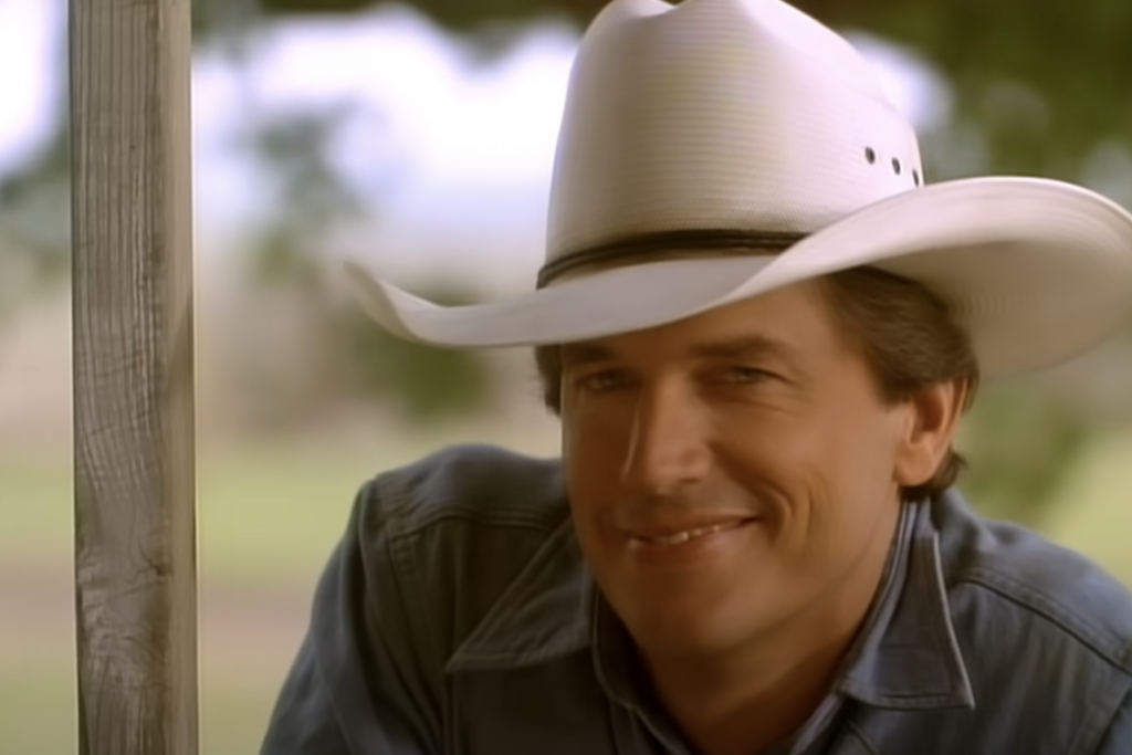 A person is smiling warmly, wearing a white cowboy hat and a blue denim shirt, leaning slightly against a wooden post. The background is blurred greenery, suggesting an outdoor setting.