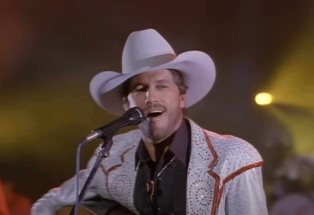 A man in a white cowboy hat and ornate jacket sings into a microphone. The background is softly lit with yellow lights, creating a warm ambiance. He appears to be performing on stage, with a focused and passionate expression.