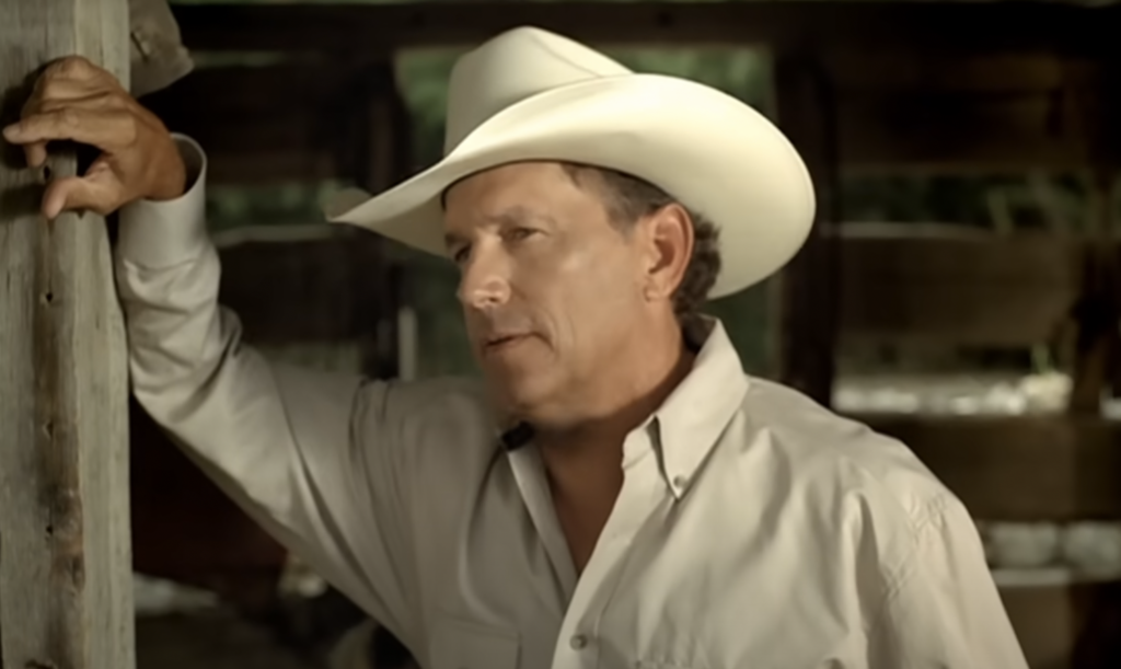 A man in a white cowboy hat and cream-colored button-down shirt leans against a wooden post. He is outdoors, possibly in a rustic, barn-like setting, with a contemplative expression on his face.