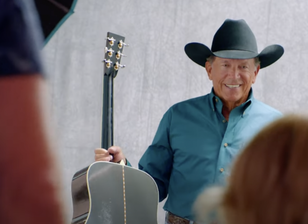 A man wearing a cowboy hat and a blue shirt smiles while holding a guitar. He is standing in front of a neutral background. A blurred figure is visible in the foreground on the right side of the image.