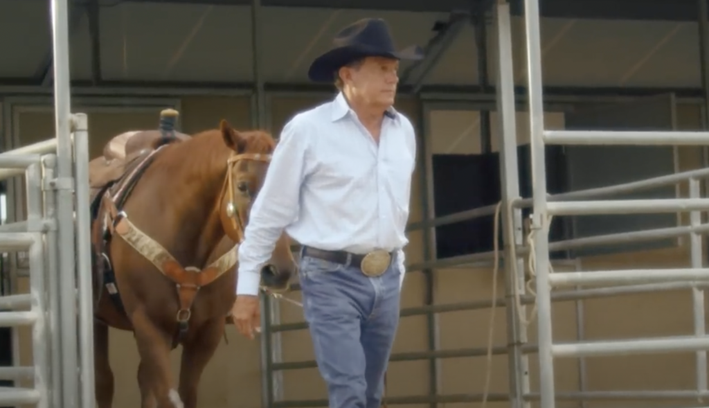 A man wearing a black cowboy hat, white shirt, and jeans walks through a metal gate leading a brown horse. A saddle is secured on the horse's back. The background features a building with large windows.