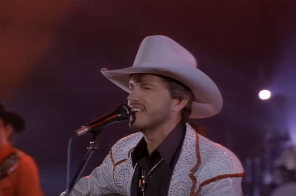 A man wearing a white cowboy hat and a white jacket with red accents sings into a microphone on stage. He has facial hair and appears to be performing country music. The background is dimly lit with colorful stage lights. Another musician is partially visible.