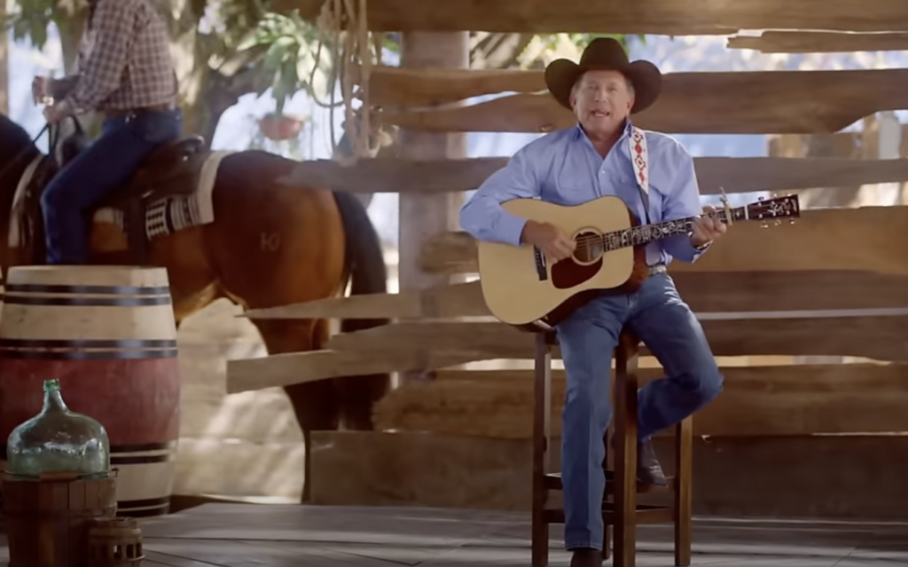 A man wearing a cowboy hat and blue shirt sits on a wooden stool, playing an acoustic guitar and singing. Behind him, another person on horseback is partially visible. The setting appears to be a rustic, wooden outdoor area with barrels and a large glass jug.