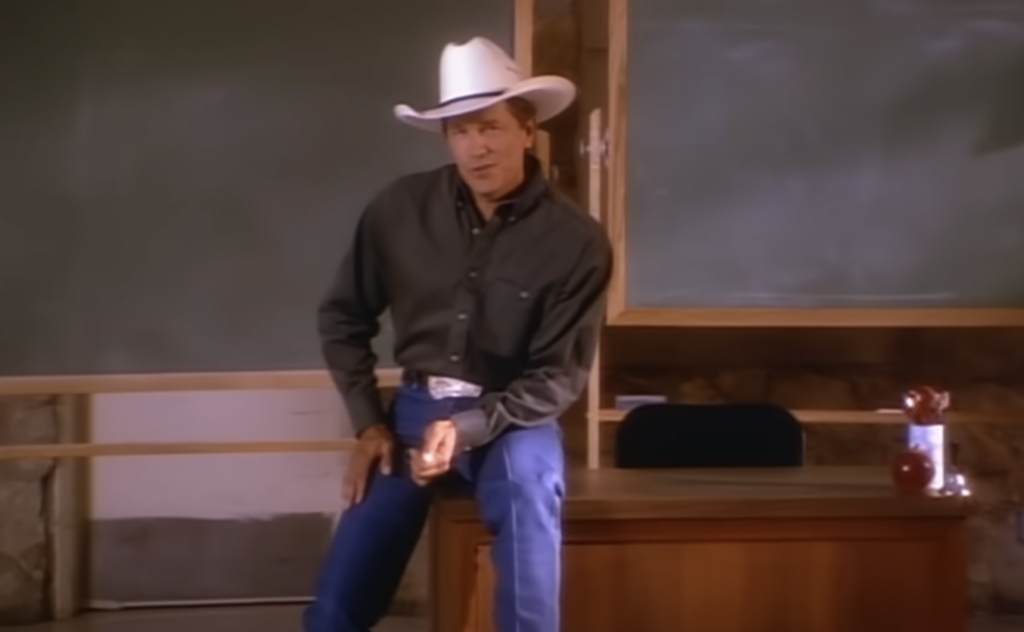 A man wearing a black shirt, blue jeans, and a white cowboy hat leans against a wooden desk in front of a blackboard. An apple is placed on the desk. The setting resembles a classroom.