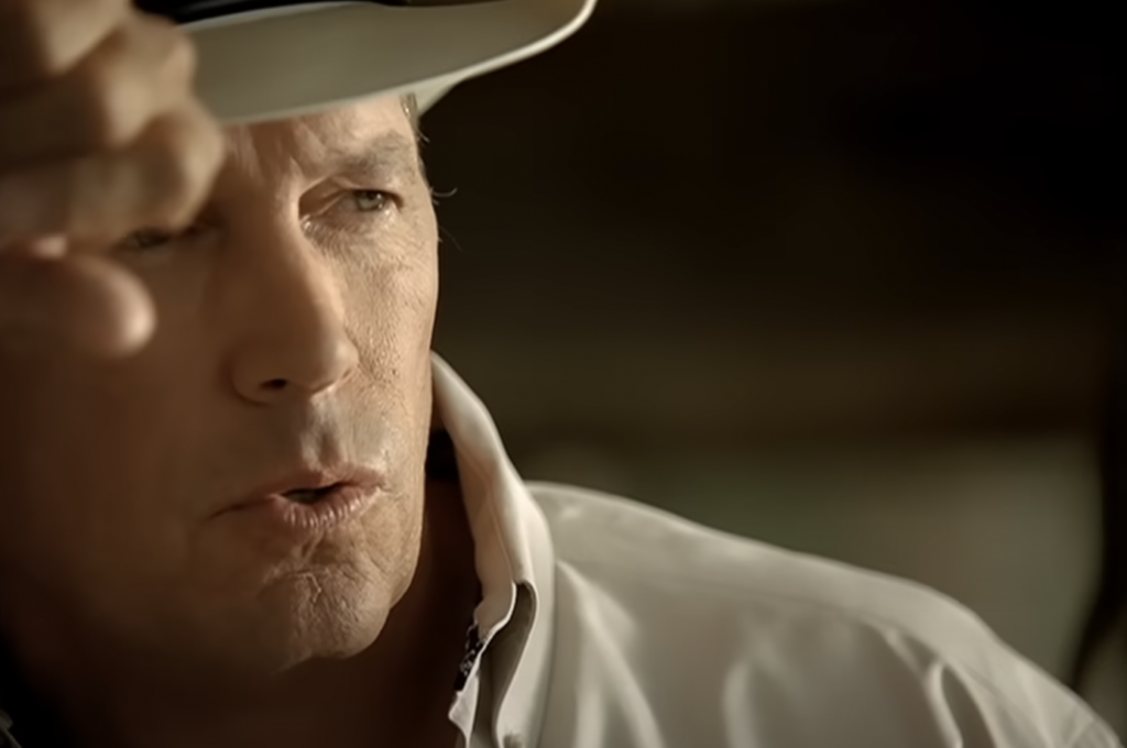 Close-up of a person wearing a white shirt and a white hat, depicted partially in shadow. The individual is touching the brim of their hat and appears to be in a thoughtful or intense moment, looking slightly to the side. The background is blurred and indistinct.