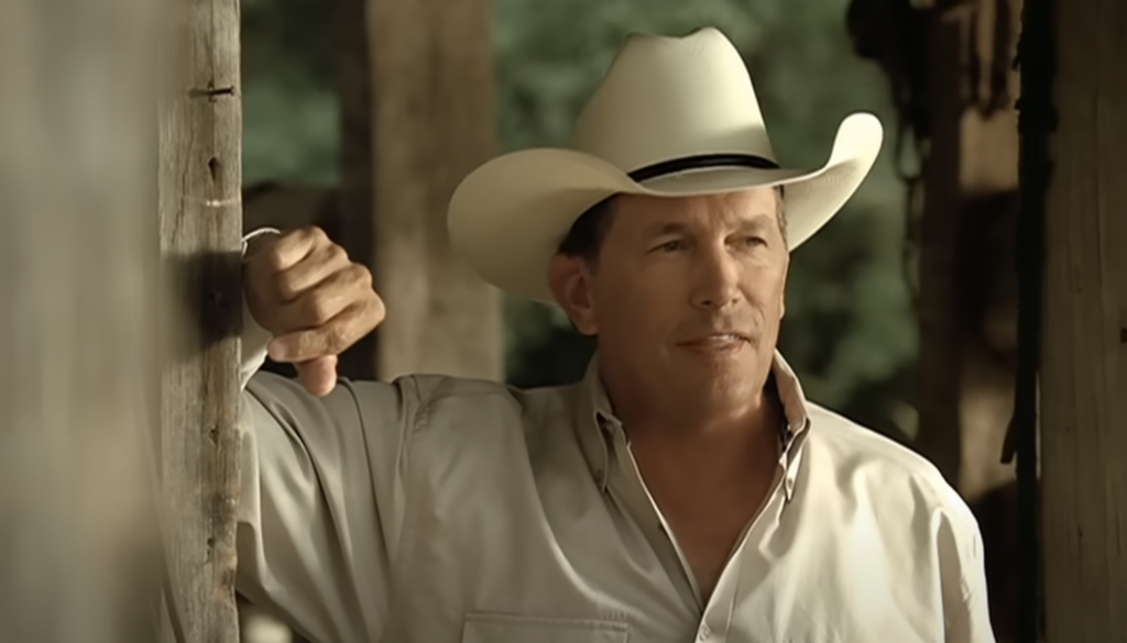 A man wearing a white cowboy hat and a light-colored button-up shirt leans against a rustic wooden post, looking off to the side. He appears to be outdoors with a blurry, nature-filled background.