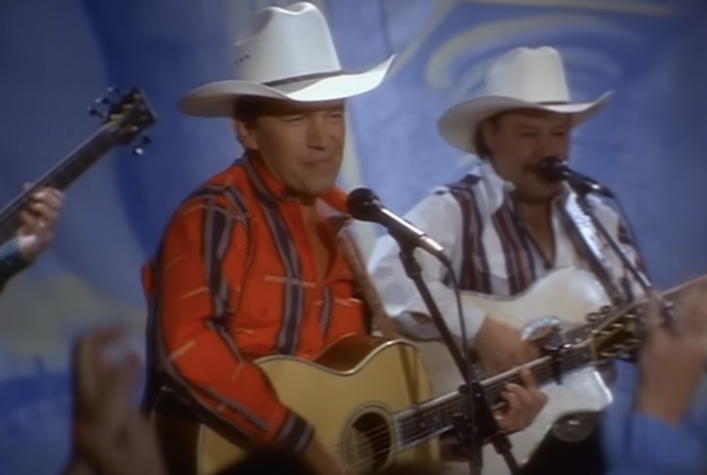 Two men wearing white cowboy hats perform on stage, each playing a guitar and singing into microphones. The man in the foreground wears a red shirt with black stripes, and the man in the background wears a striped shirt. A blurred audience member can be seen in the foreground.