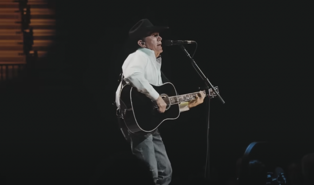 A musician wearing a cowboy hat and a light-colored shirt performs on stage, singing into a microphone and playing a black acoustic guitar. The background is dark, featuring some lights and a small portion of the audience visible in the foreground.