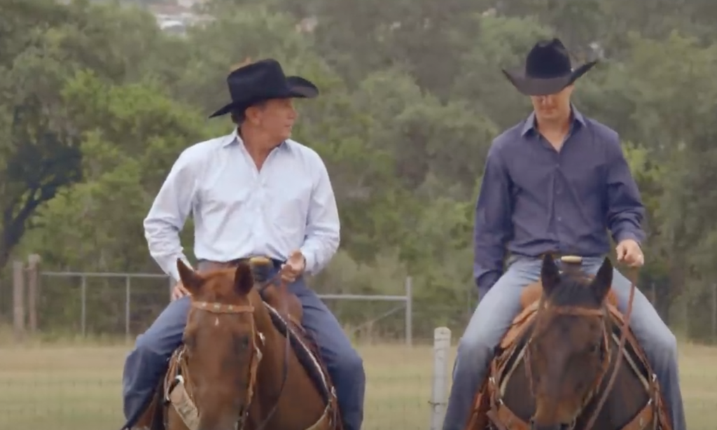 Two people are riding horses side by side in an outdoor setting. They are both wearing cowboy hats and long-sleeved shirts. The background shows a fenced field with trees. The person on the left is looking at the person on the right, while the latter gazes forward.