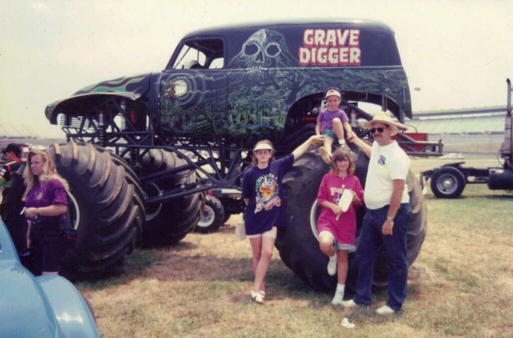 A group of people, including two adults and two children, stand beside a large monster truck called "Grave Digger" with detailed artwork on its body. One child is sitting on the truck's huge tires. The scene appears to be at an outdoor event on a sunny day.