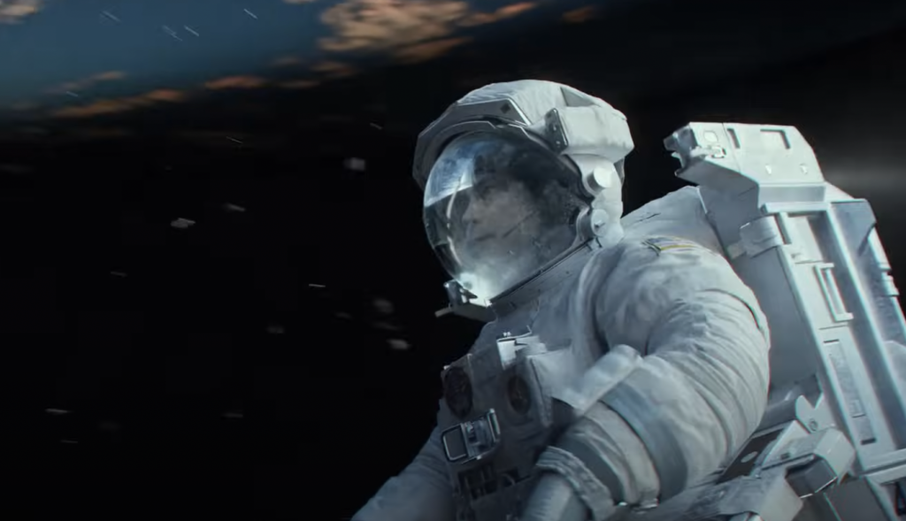 An astronaut in a spacesuit appears to be floating in outer space, with a dark backdrop and faint glowing clouds visible in the distance. The astronaut's helmet visor shows reflections of light.