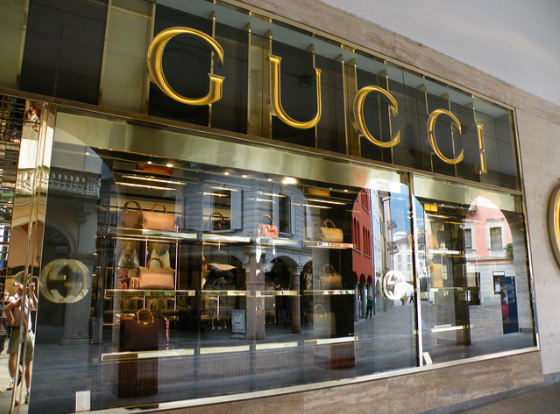 A storefront of a Gucci retail store with large glass windows displaying handbags and accessories. The store's name "Gucci" is prominently featured in large gold letters above the entrance. Reflections of buildings and a street scene are visible in the glass.