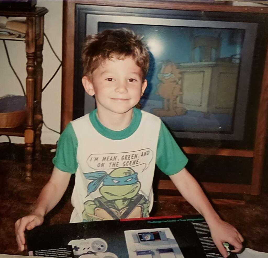 A young boy with short brown hair smiles while kneeling on the carpet, holding a boxed Super Nintendo Entertainment System. He is wearing a Teenage Mutant Ninja Turtles shirt. In the background, a TV displays a scene from a Garfield cartoon.