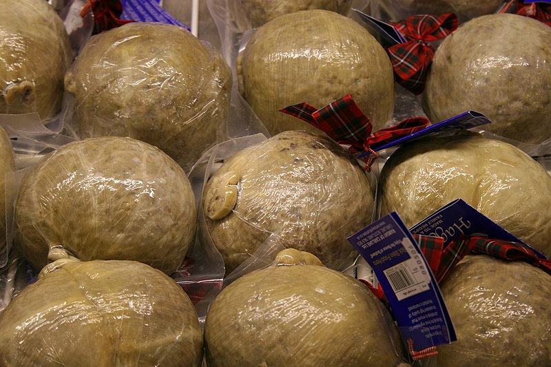 A display of several haggis, wrapped in plastic and tied with red tartan ribbons. The haggis are round and beige in color, and each has a label attached. They are tightly packed together, occupying most of the image.