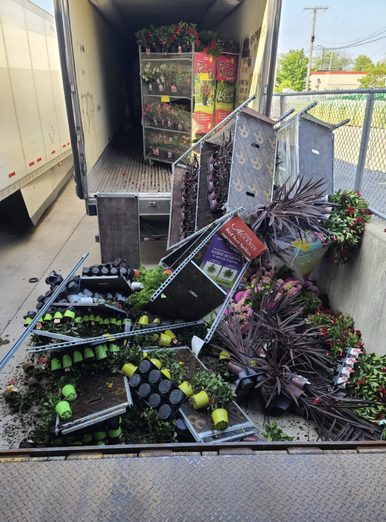A variety of plants and flower pots spilled out from an open truck trailer, causing a mess near the truck dock. The fallen plants and pots are scattered on the ground, with some shelving units also toppled over.
