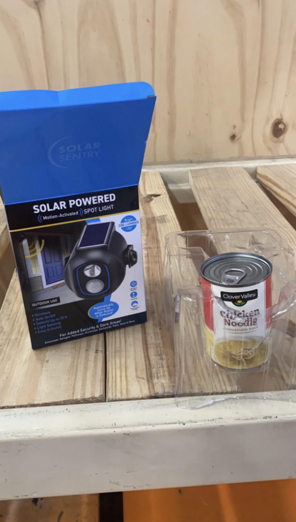 A boxed Solar Sentry solar-powered, motion-activated spotlight and a can of Clover Valley chicken noodle soup are placed on a wooden palette. The can is in a clear plastic holder beside the box.