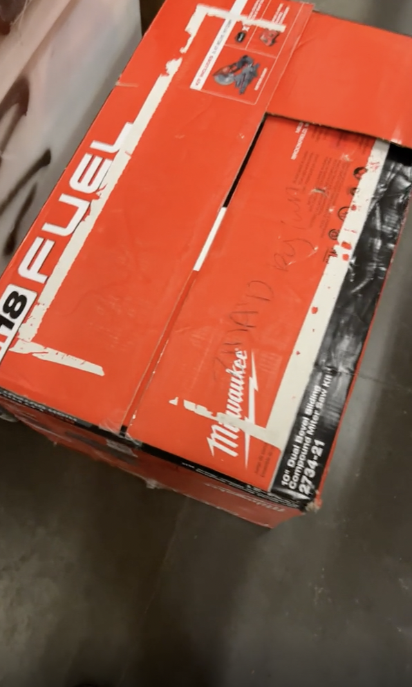 A red and brown cardboard box labeled "Milwaukee M18 FUEL" is partially open. The box appears to be for a power tool. There is handwritten text on the top of the box, and it is placed on a tiled floor next to another packaged item.