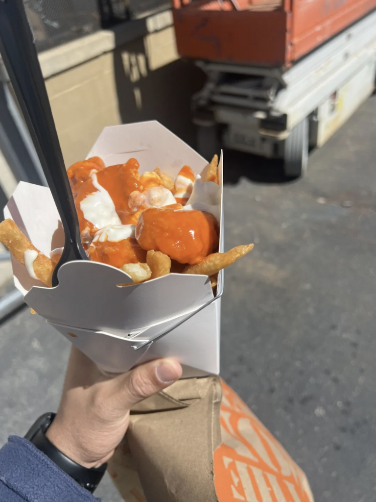 A hand holding a white container filled with fries topped with orange and white sauces. The container is held above a brown paper bag, and a black plastic fork is inserted into the fries. The background has outdoor construction equipment and pavement.