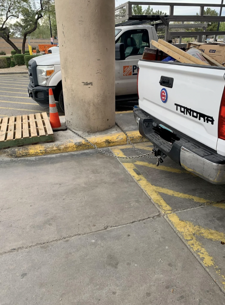 A white Toyota Tundra pickup truck is parked with its rear chained to a large concrete column in a parking lot. A wooden pallet lies near the back of the truck. Traffic cones are positioned near the truck and a nearby white utility vehicle.